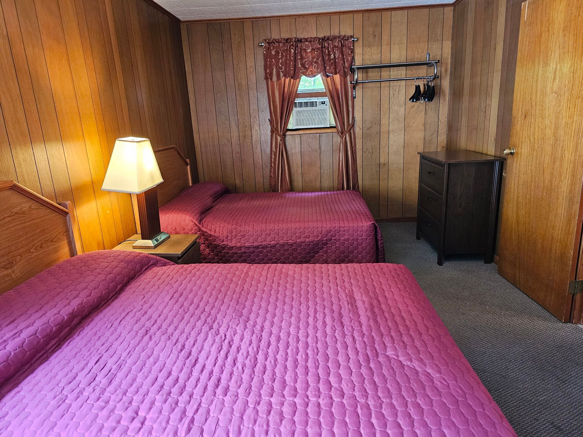 Small wood-paneled bedroom with two pink beds, a lamp, window, and dresser