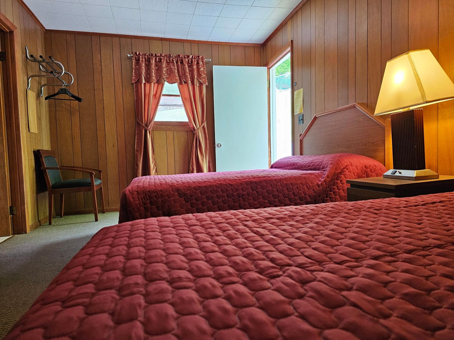 Wood-paneled motel room with two red beds, a lamp, chair, and open window curtains