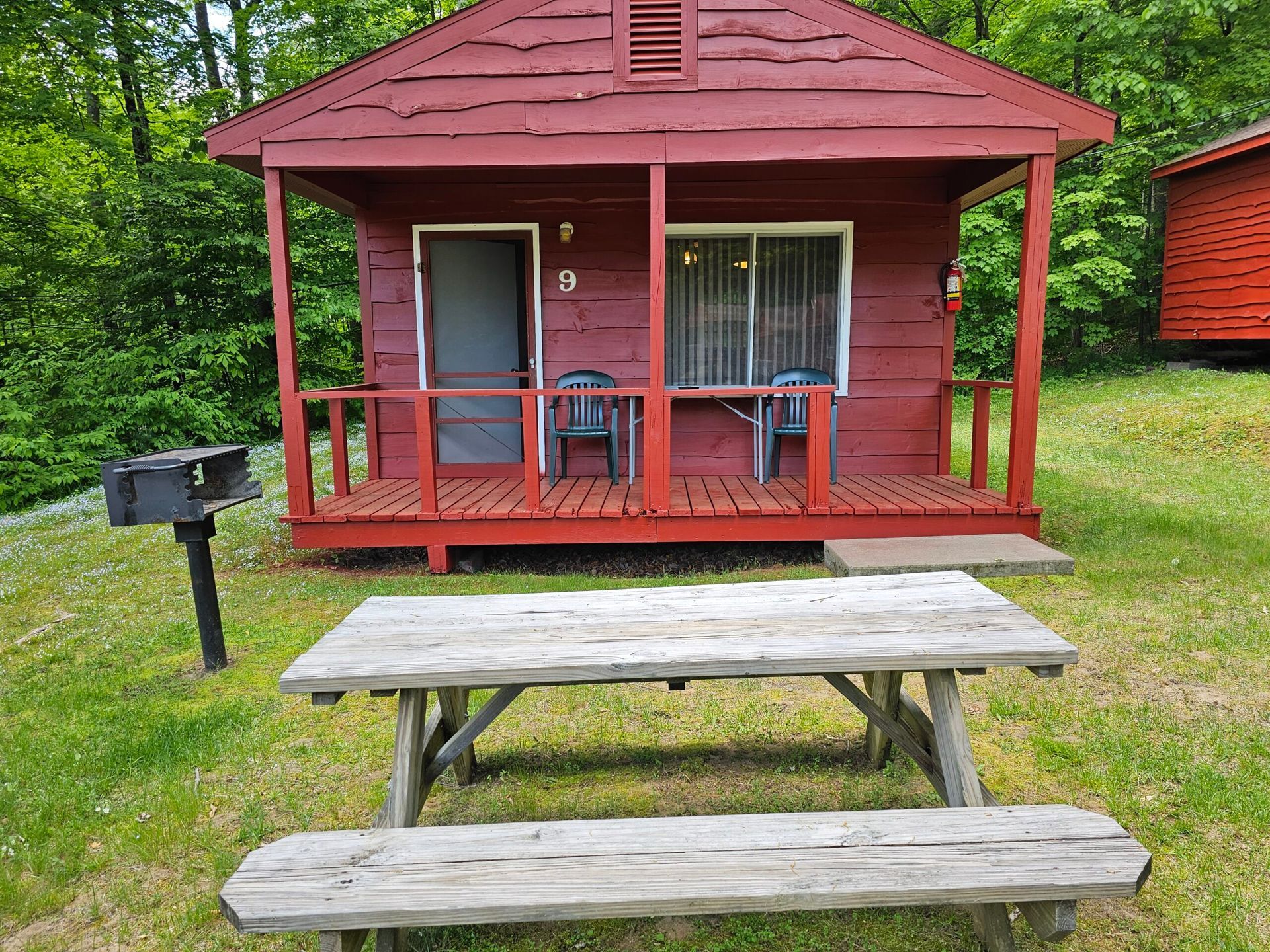 Red cabin porch with picnic table in front and a grill on the left in a grassy yard