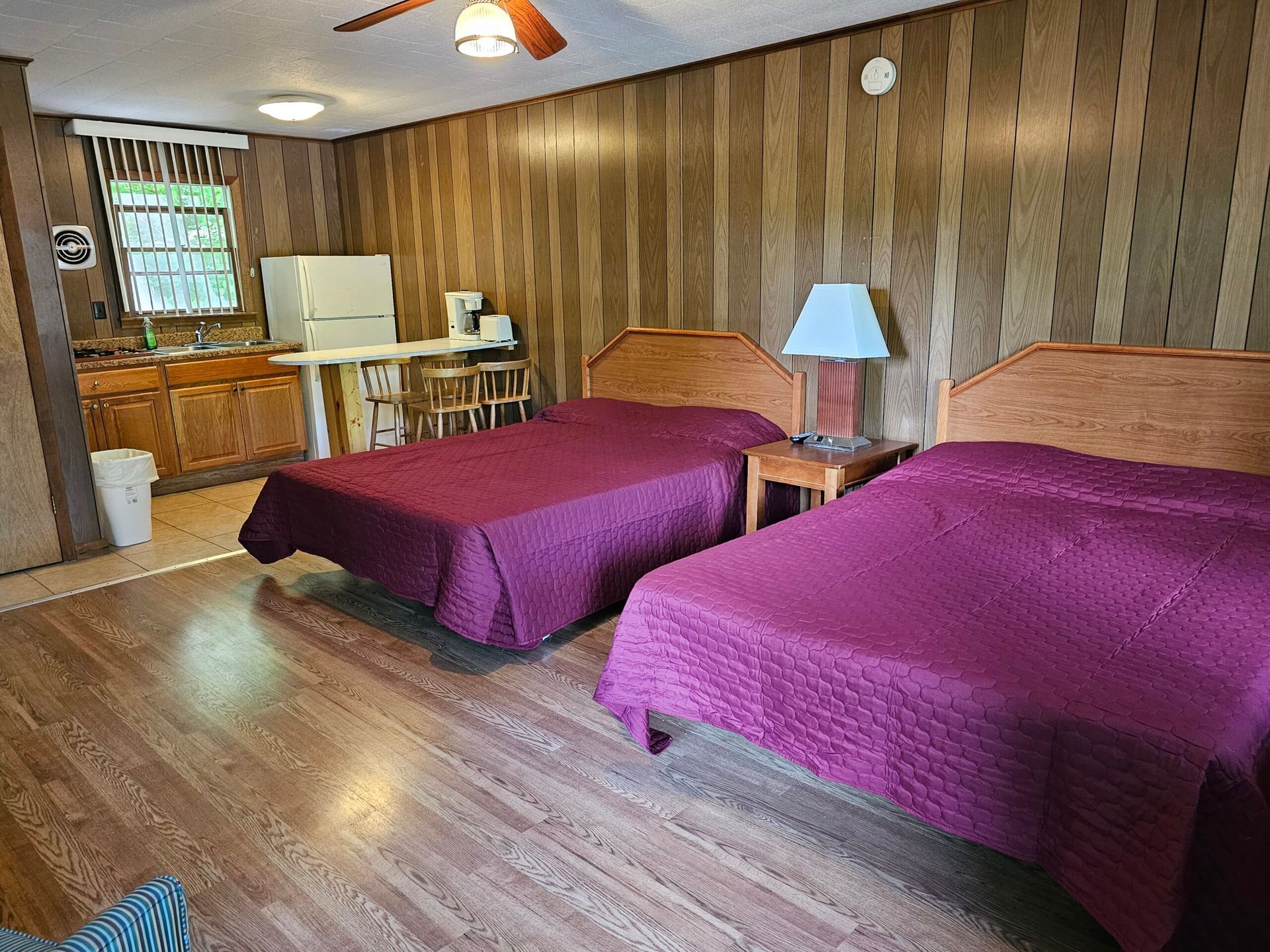 Motel room with two beds, wood-paneled walls, a lamp, and a small kitchenette area