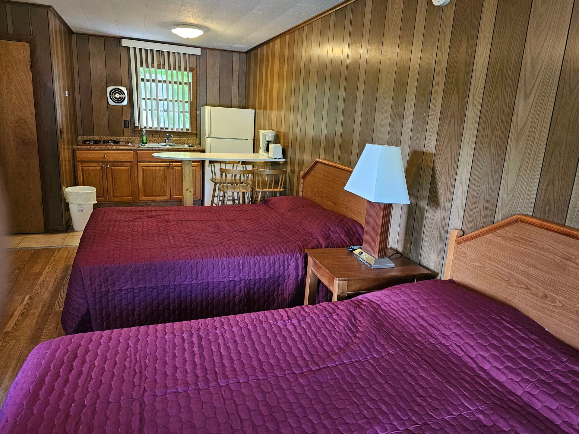 Hotel room with two purple beds, wood-paneled walls, a lamp, and a small kitchenette area