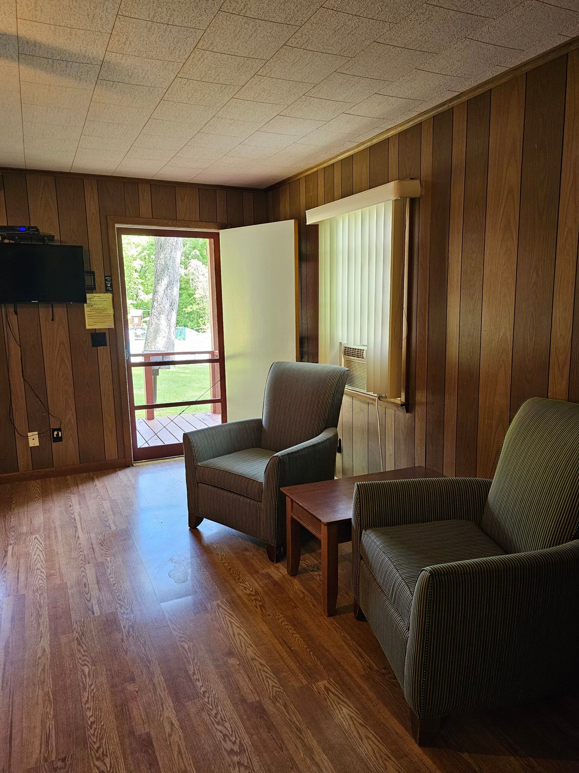 Wood-paneled living room with two striped armchairs, a window, and a doorway letting in daylight.