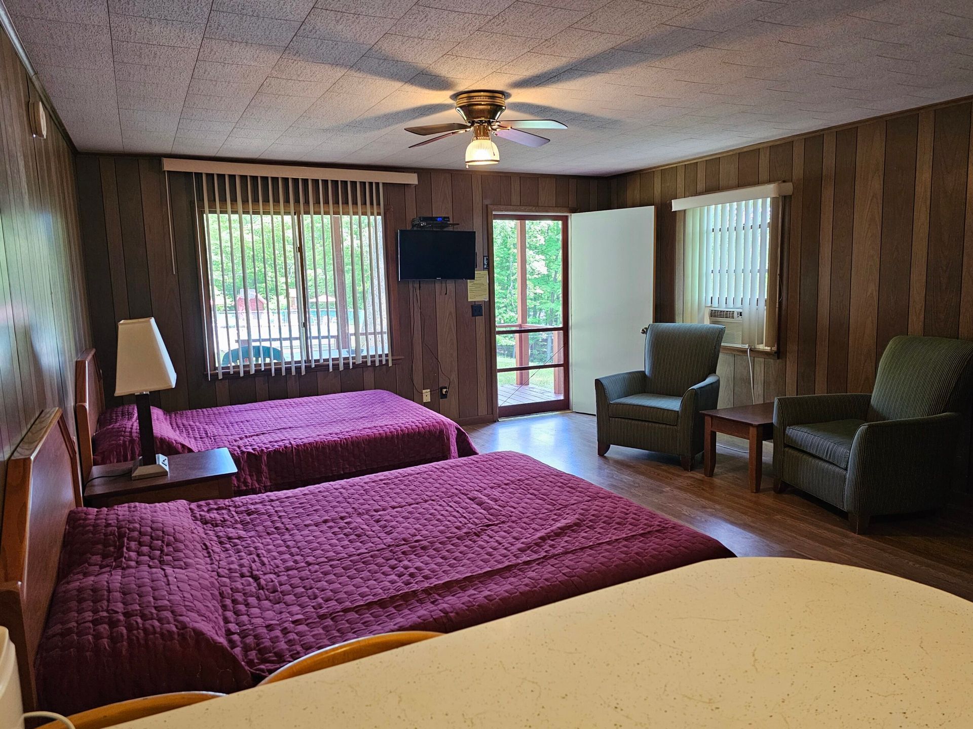 Motel room with two purple beds, armchairs, TV, ceiling fan, and sliding glass door to a balcony.