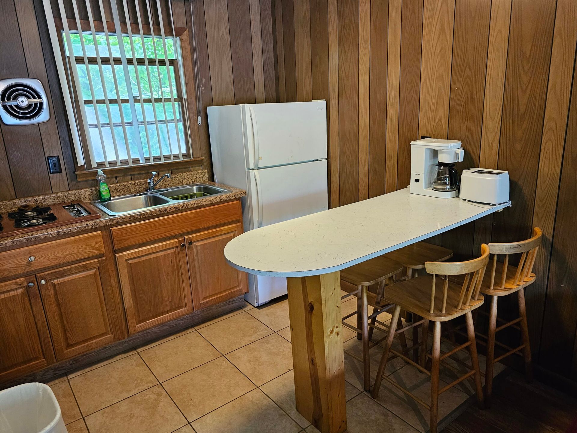 Kitchen with wood-paneled walls, white fridge, sink, and a curved breakfast bar with stools and coffee maker.