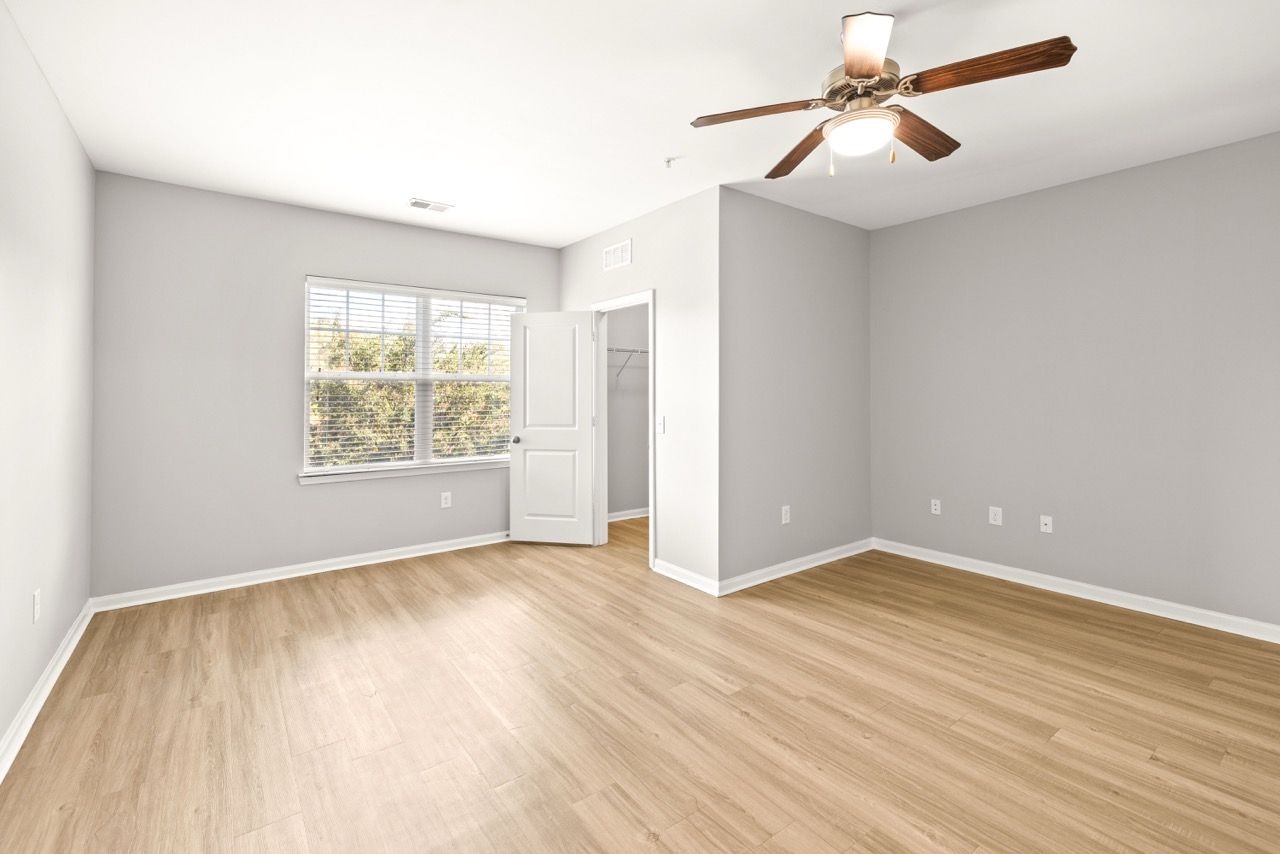 Empty gray apartment bedroom with wood-like floors, a window, ceiling fan, and an open closet door.
