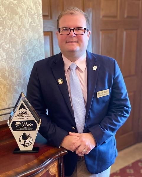 A person in a blue blazer and light blue tie stands beside a 2020 award plaque in a patterned room.