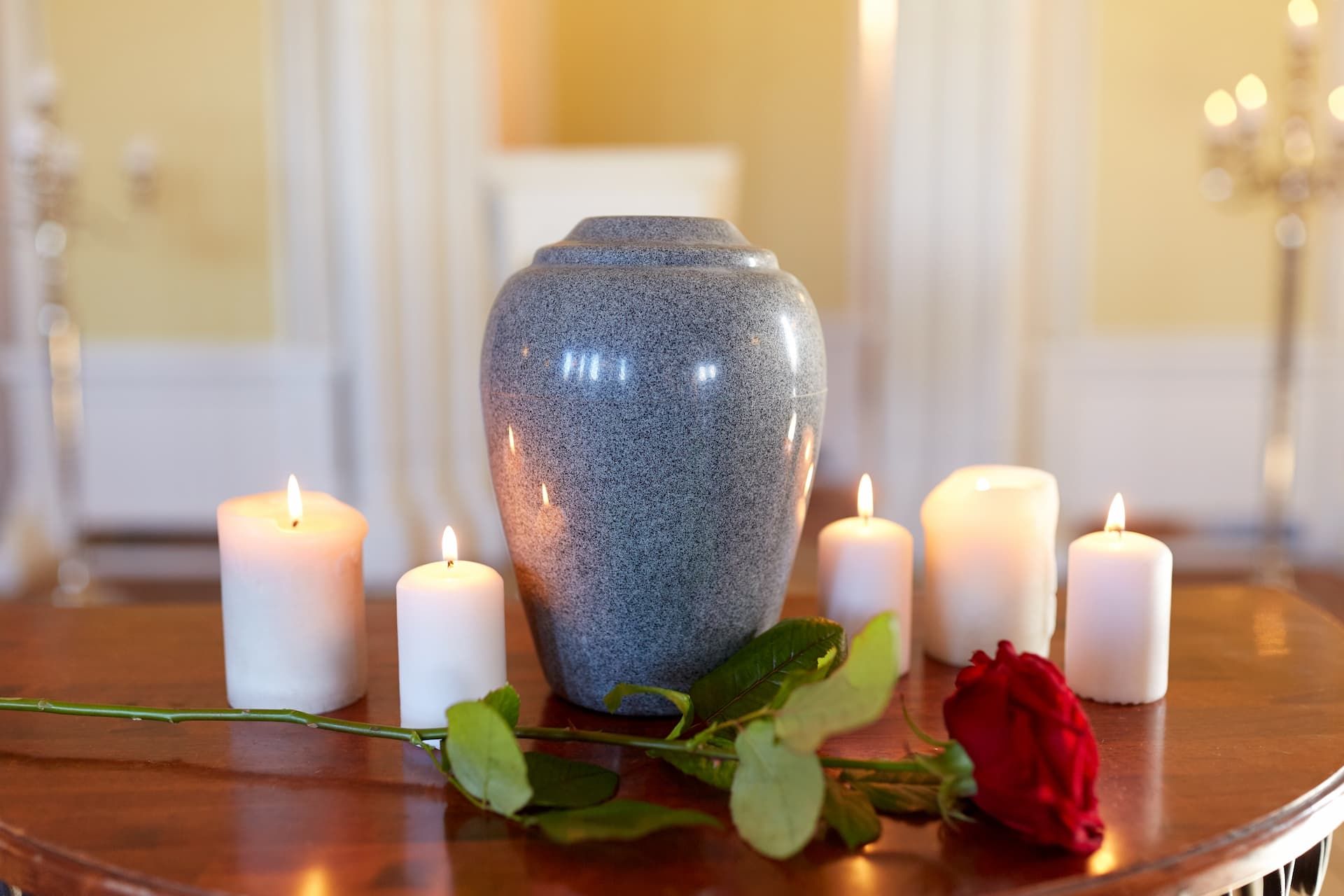 A grey stone urn sits on a wooden table, surrounded by lit candles and a single red rose in a softly lit room.