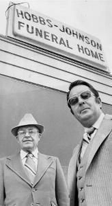 Two men in suits and ties stand in front of a sign reading 