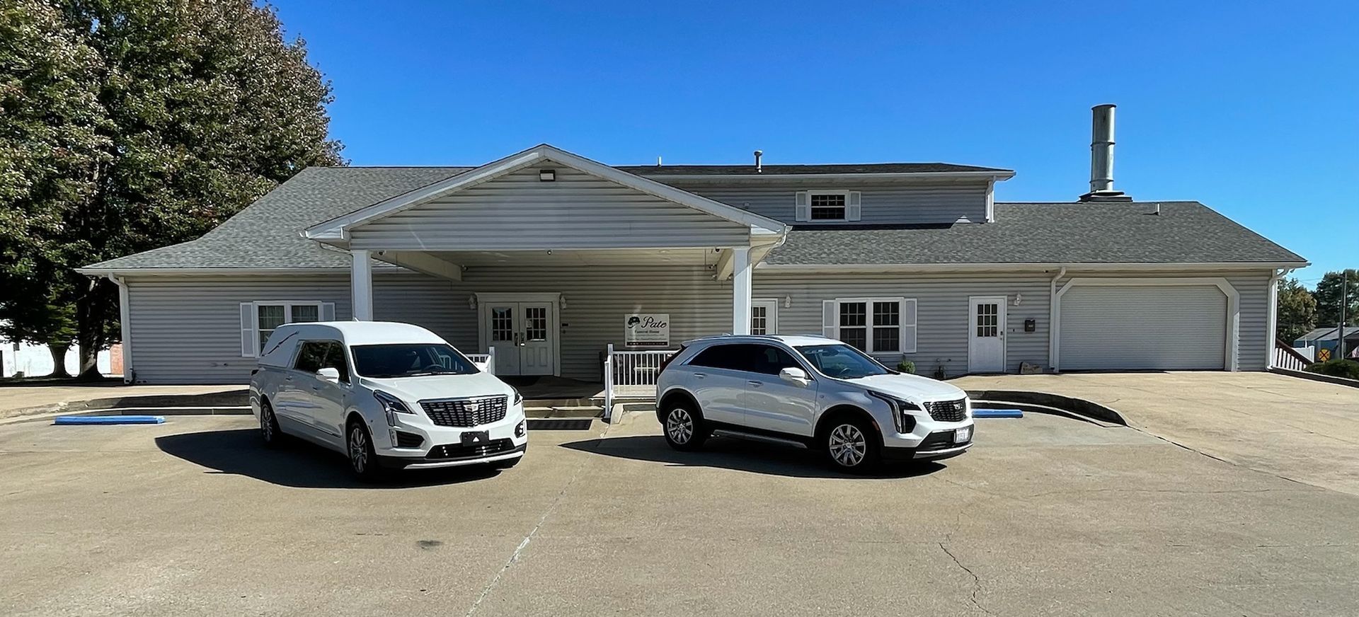 Two white funeral home vehicles parked in front of a light gray single-story building with a covered entrance.