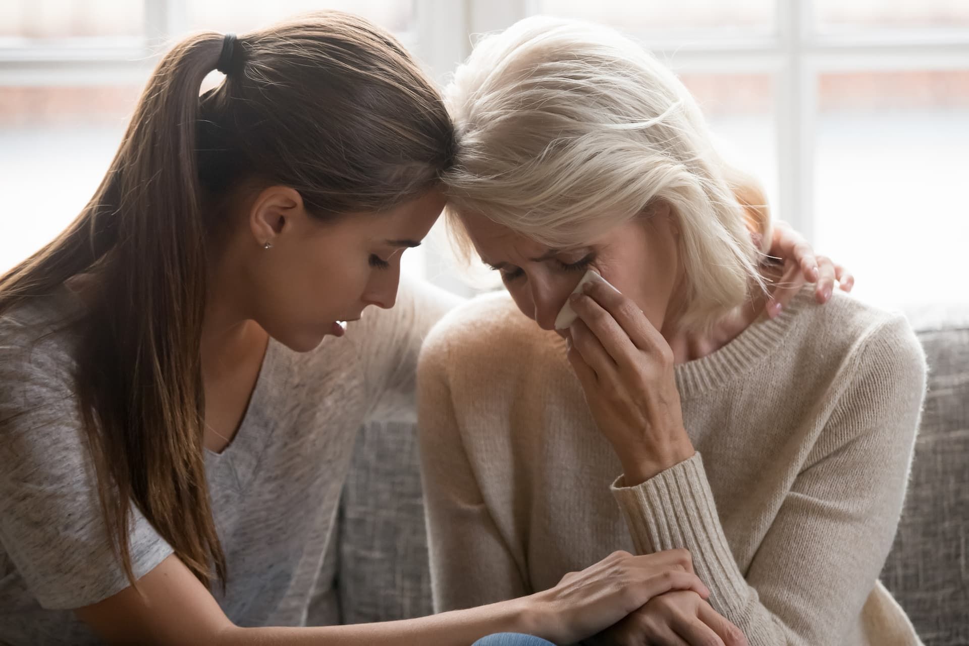 A young person comforts a crying adult, resting their forehead against hers while holding her hand.