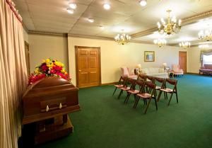 A wooden casket topped with colorful flowers sits in a funeral home visitation room with green carpet and rows of chairs.