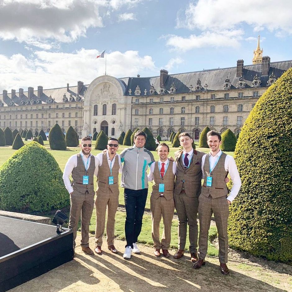 Five men posing in front of a historic building and manicured hedges on a sunny day