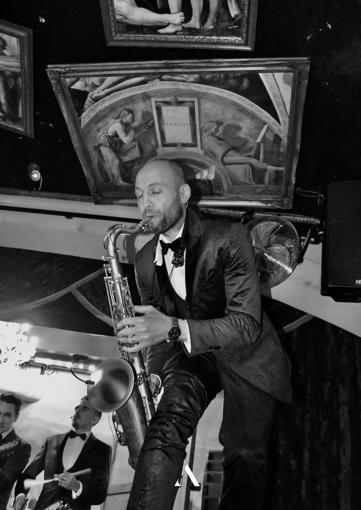 Saxophonist performing onstage in a suit, with a drummer visible in the background.