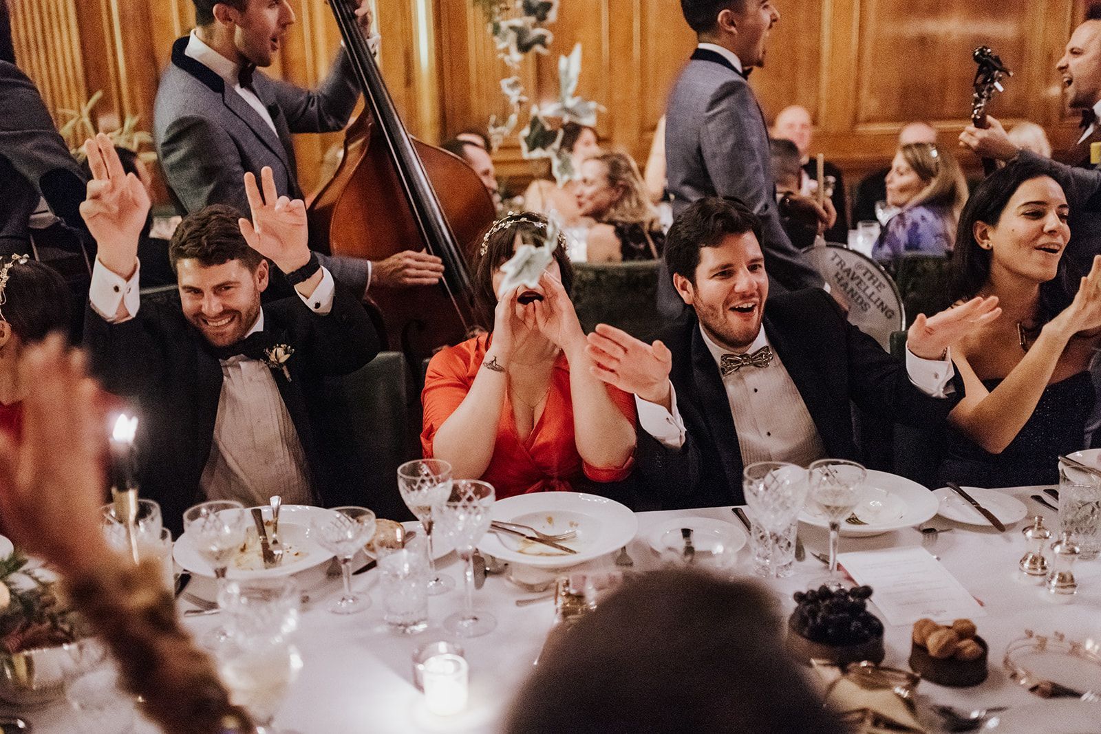 Guests cheer and raise hands at a banquet table during a lively celebration, with musicians in the background