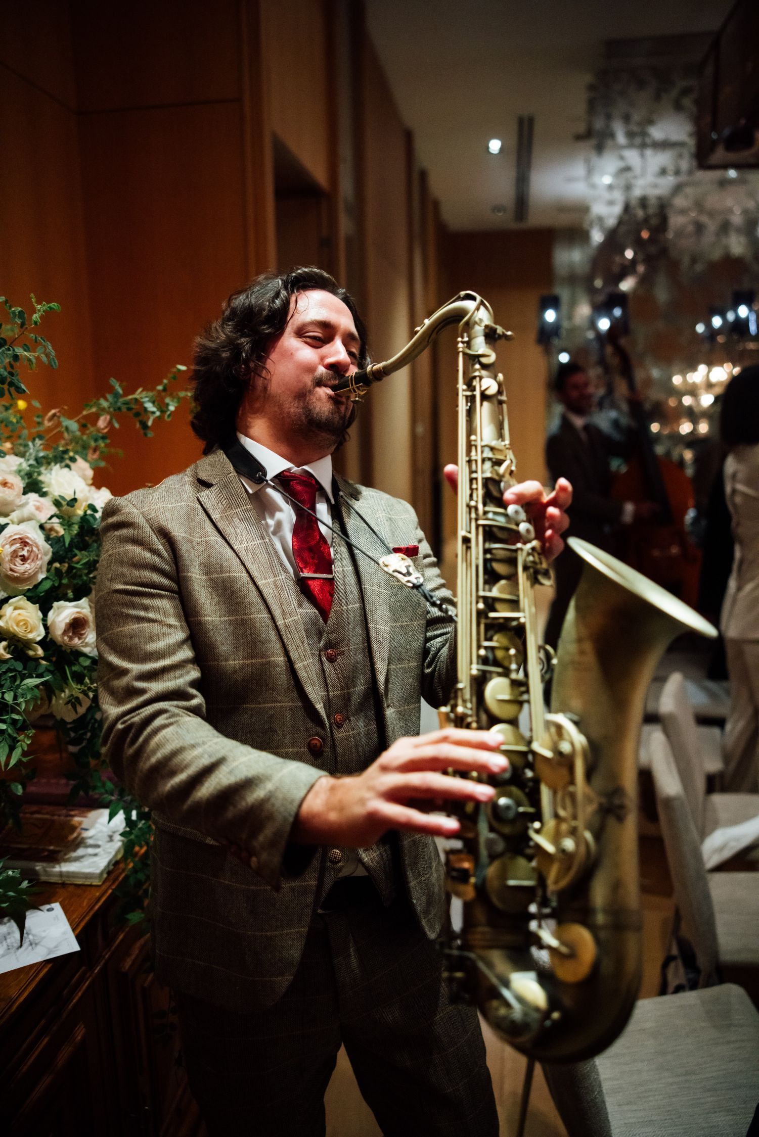 Musician playing a saxophone in a warmly lit indoor venue with flowers and reflections nearby