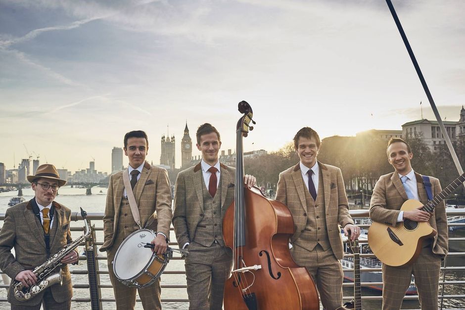 Five musicians in matching suits perform outdoors by a waterfront at sunset.