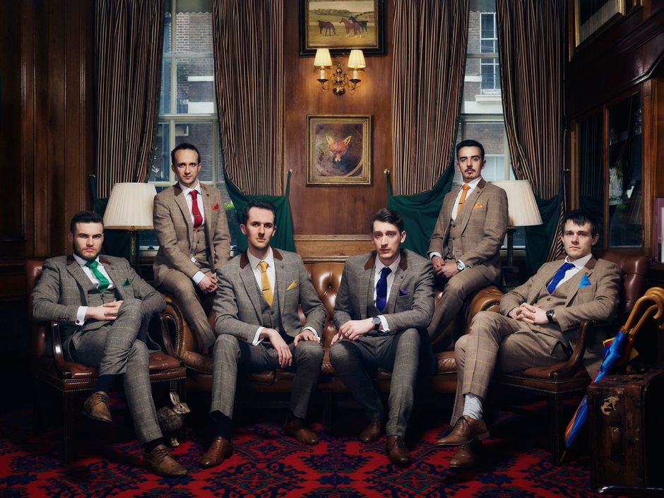 Six men in suits seated in a wood-paneled lounge, posing formally with vintage decor and lamps behind them.