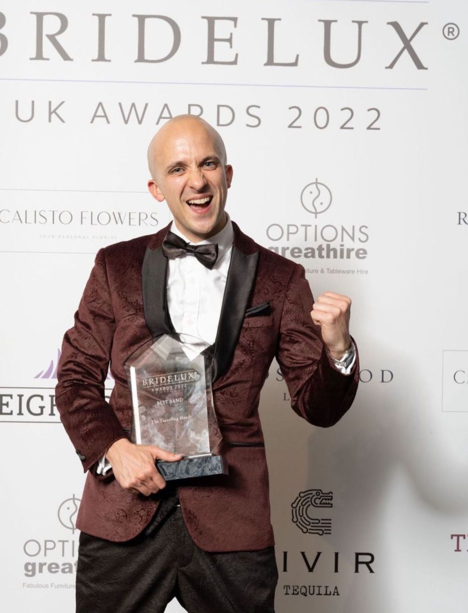 Man in burgundy tuxedo holding an award and smiling at the BRIDELUX UK Awards 2022 backdrop