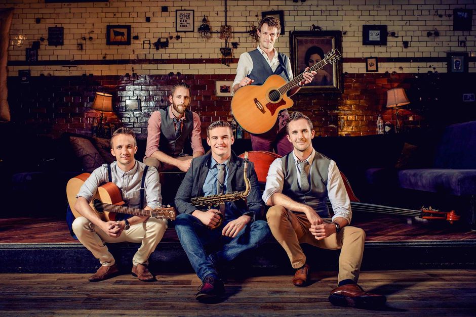 Five musicians in a rustic venue, one standing with an acoustic guitar, four seated in front