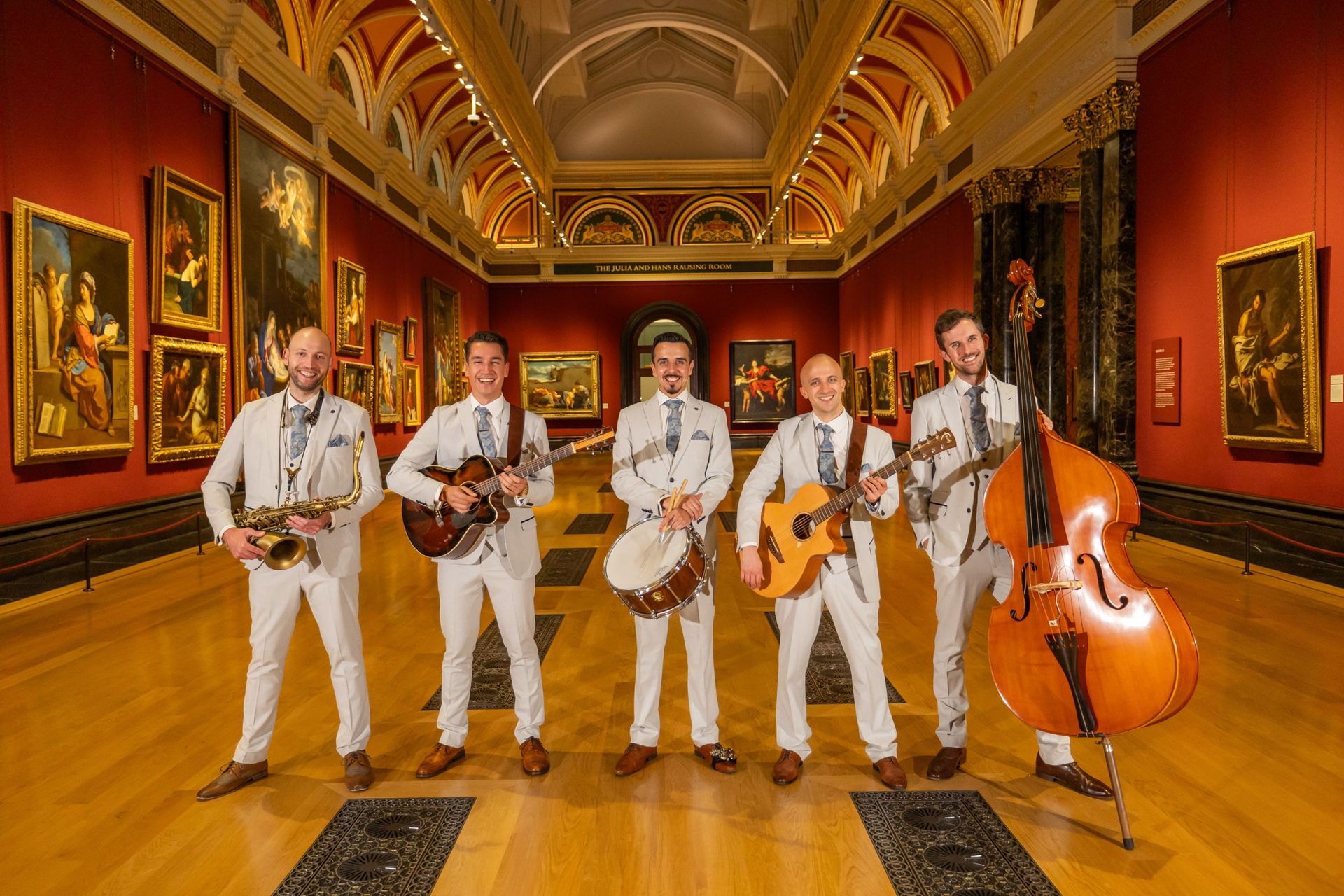Five musicians in white suits performing in a grand ornate room with gold-framed paintings and a cello.