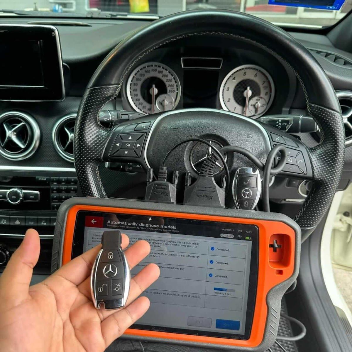 A person holding a Mercedes key, with a diagnostic tool and car's steering wheel in the background.