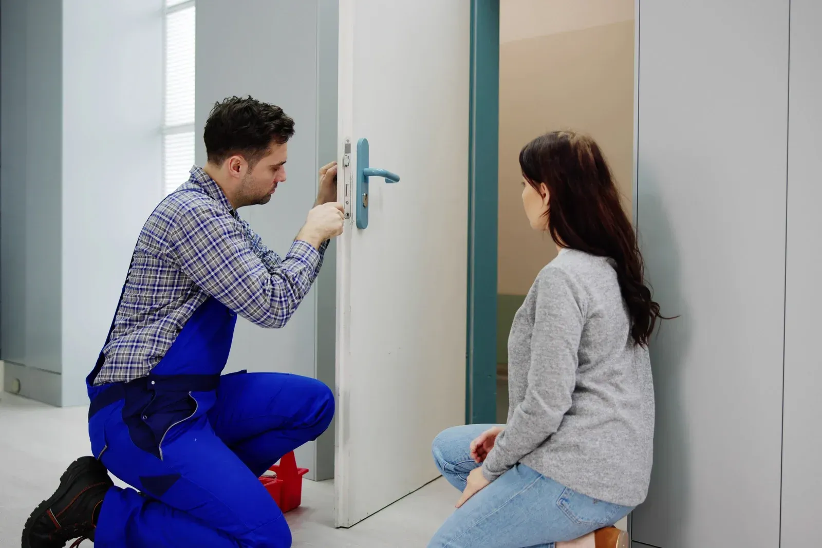 A locksmith in blue workwear kneels to repair a door lock while a person watches, in a bright, modern room.