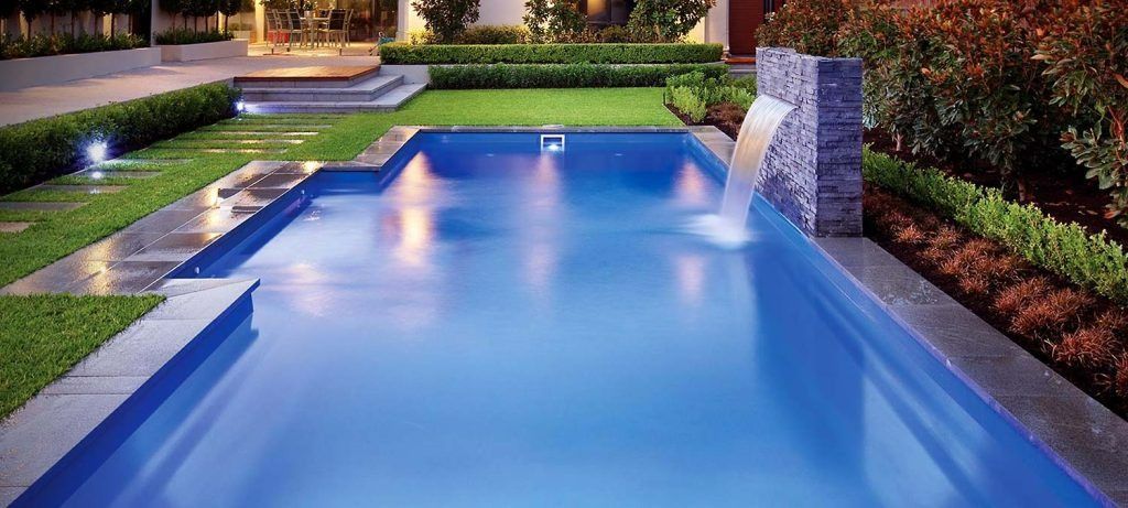 A rectangular backyard swimming pool with a stone waterfall feature, surrounded by manicured lawn and garden beds.