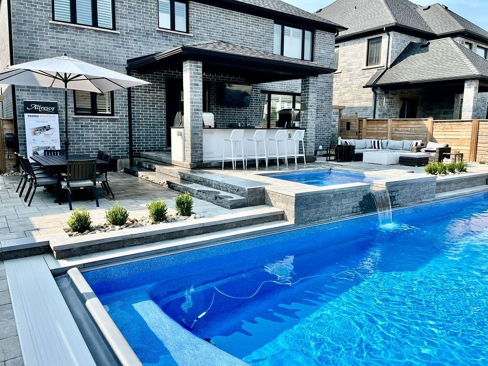 A sleek indoor swimming pool with blue water, floor-to-ceiling windows, and orange lounge chairs on a stone deck.