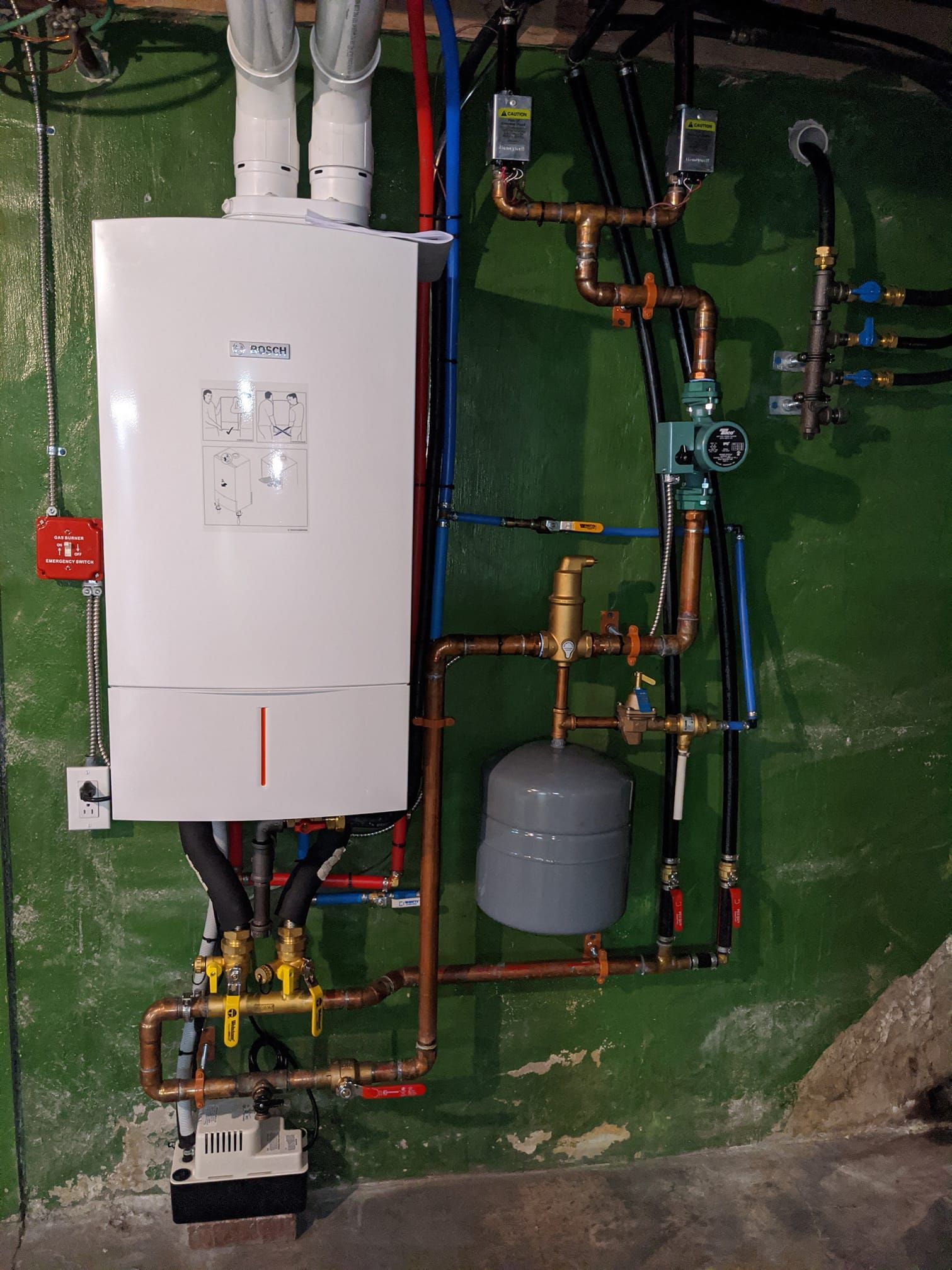 A white boiler is surrounded by copper pipes on a green wall