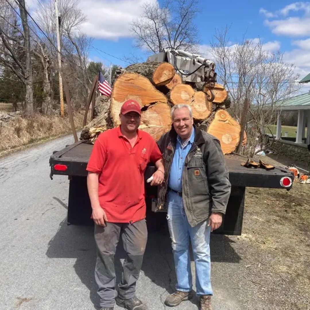 Two people standing beside a trailer loaded with cut logs on a roadside, one in a red shirt and cap, one in a jacket