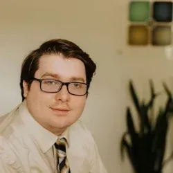 Man with glasses and tie, looking at the camera, light-colored shirt, with a plant and a wall art.