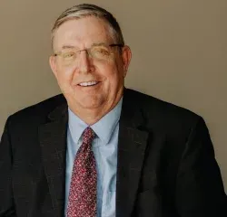 Man in suit, glasses, and tie smiling, indoor setting.