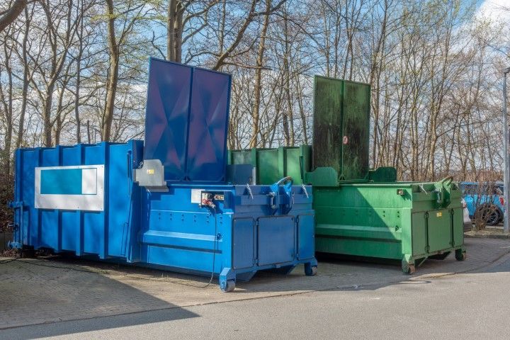 A blue and a green industrial trash compactor sit side by side in a paved lot, with trees in the background.
