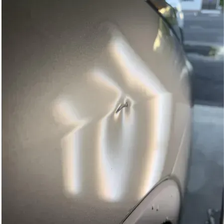 Close-up of a silver car door with a small dent and bright reflections from a nearby window