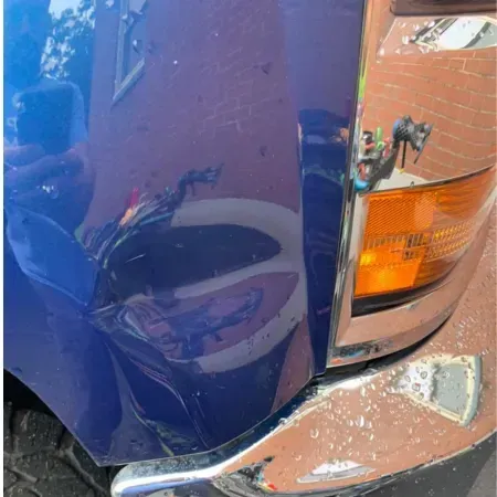 Close-up of a blue vehicle’s dented side near the orange front turn signal, with scraped silver bumper reflection.