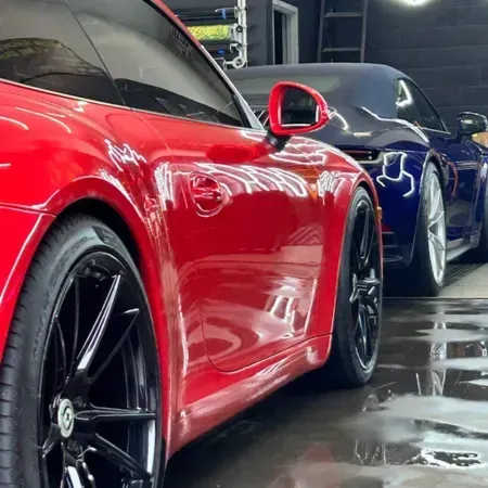 Red sports car in a garage beside a blue car, with wet floor reflections.