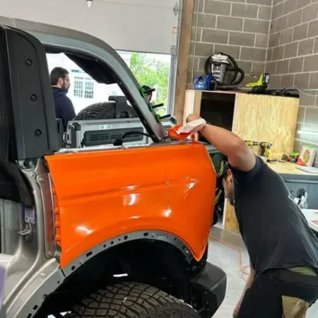 Person painting an orange vehicle fender in a garage, with another person working in the background