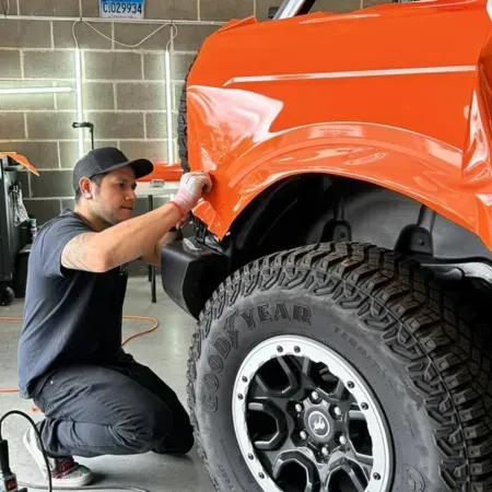 Mechanic sanding an orange truck’s fender in a garage, beside a large off-road tire.