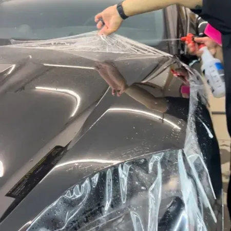 Person peeling clear protective film off a shiny black car hood in a workshop