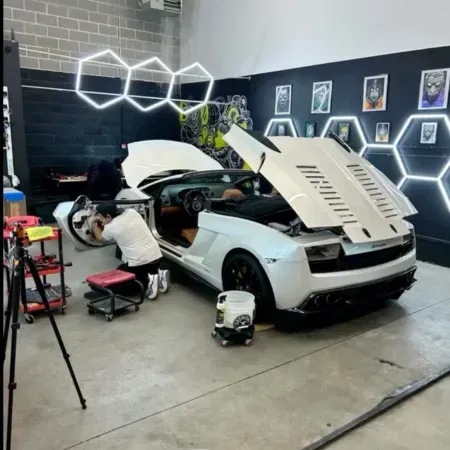Sports car in a garage with hood open, mechanic working beside it, and hexagon wall lights glowing in back.