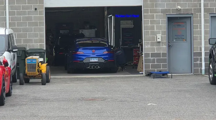 Blue sports car in an open garage bay, with tools and equipment around it and a gray building exterior.