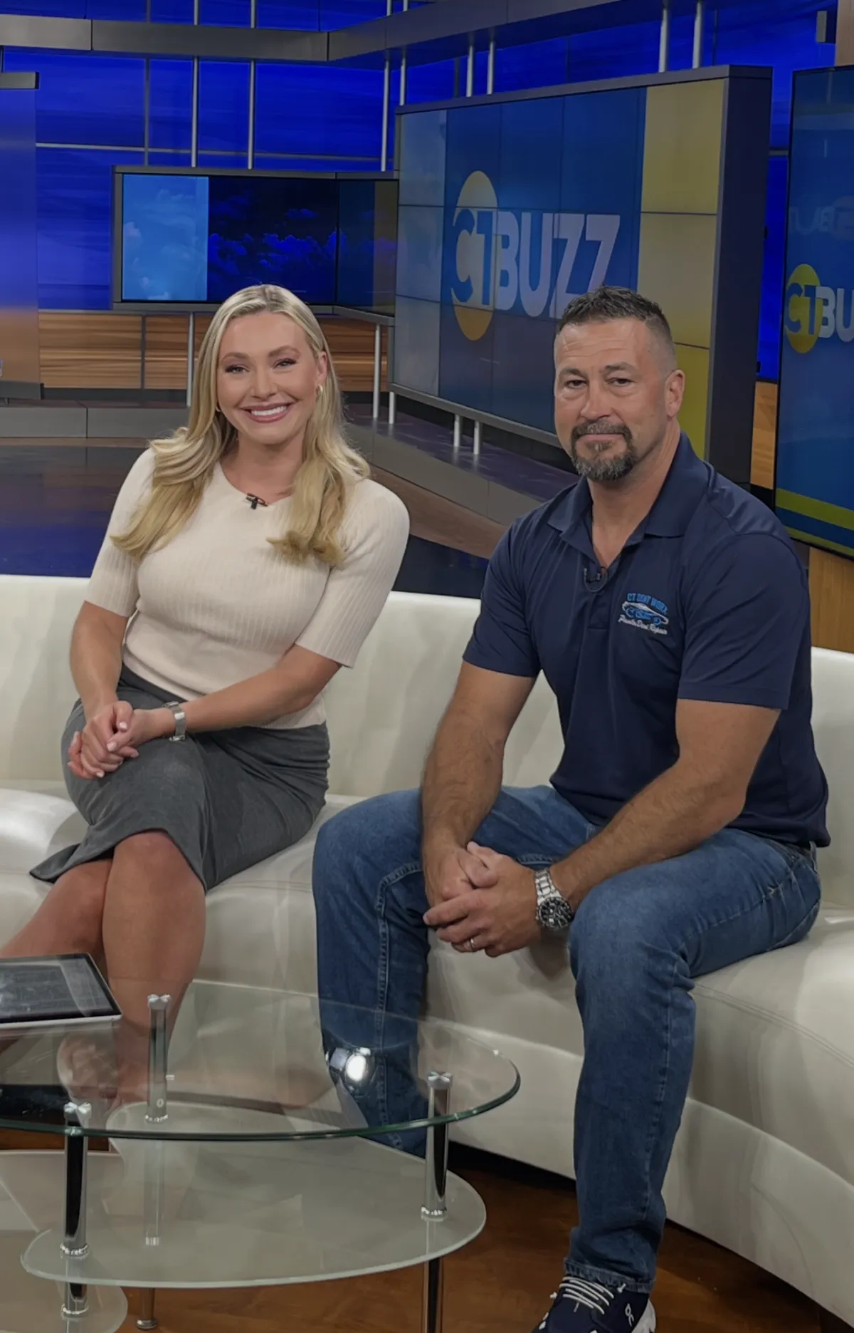 Two people seated in a TV studio with a blue “BUZZ” backdrop, smiling at the camera. Two people seated in a TV studio with a blue “BUZZ” backdrop, smiling at the camera.