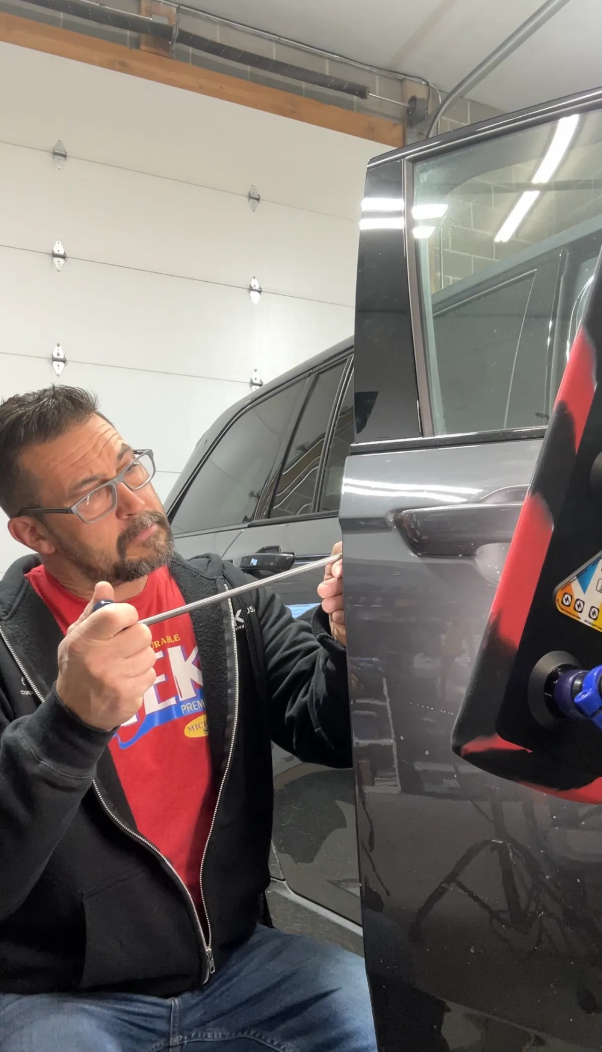 Man in glasses works on a black car in a garage, using a tool near the side panel. Man in glasses works on a black car in a garage, using a tool near the side panel.