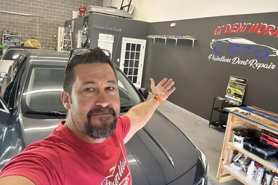 Man taking a selfie in a garage beside a black car, arm raised toward a shop sign.