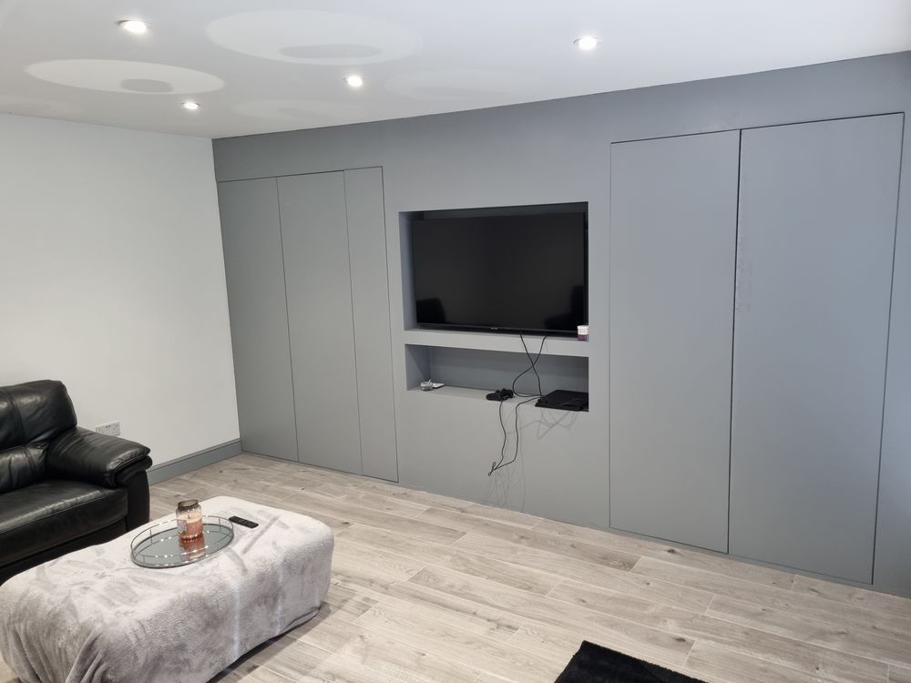 Gray-walled living room with recessed TV and storage cabinets. Gray ottoman, black sofa, and light wood-look floor.