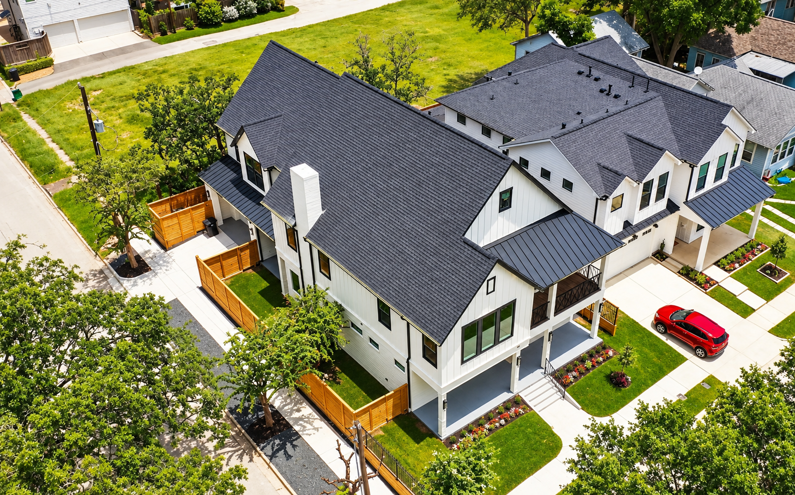 Aerial view of a large white house with dark roof, front porch, and a red car parked beside it.