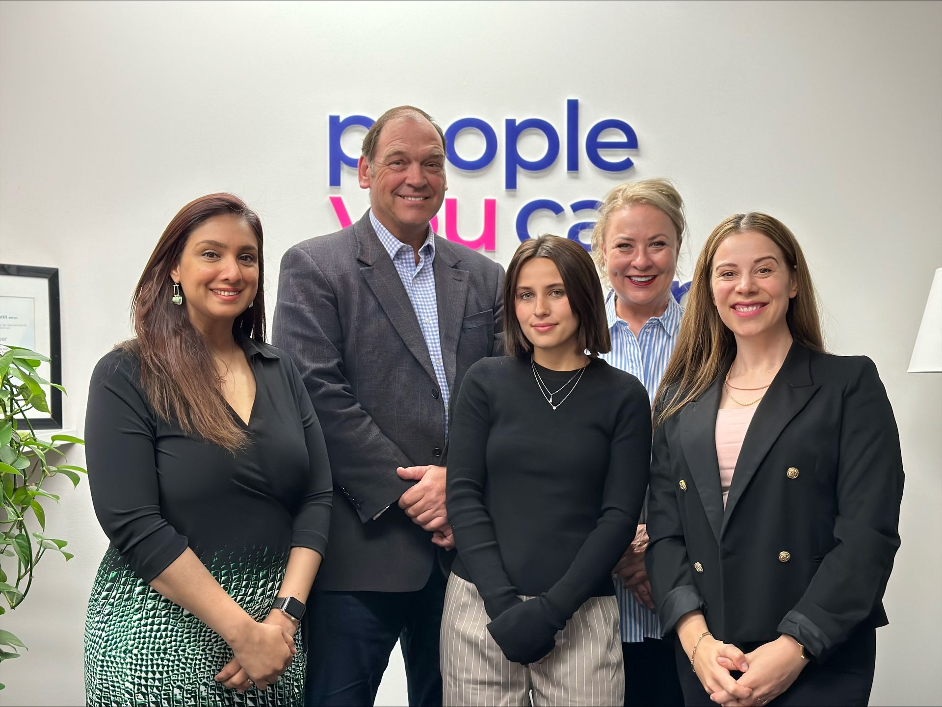 Five people smiling in an office, standing in front of a 