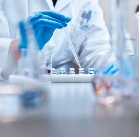 Scientist in lab coat and blue gloves uses a pipette to dispense liquid into test tubes.
