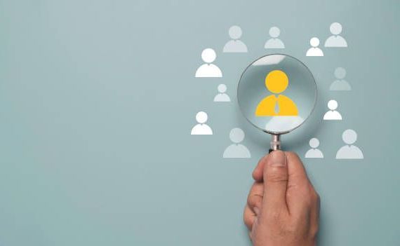 Hand holding magnifying glass focused on a yellow person icon among white icons, representing recruitment or selection.
