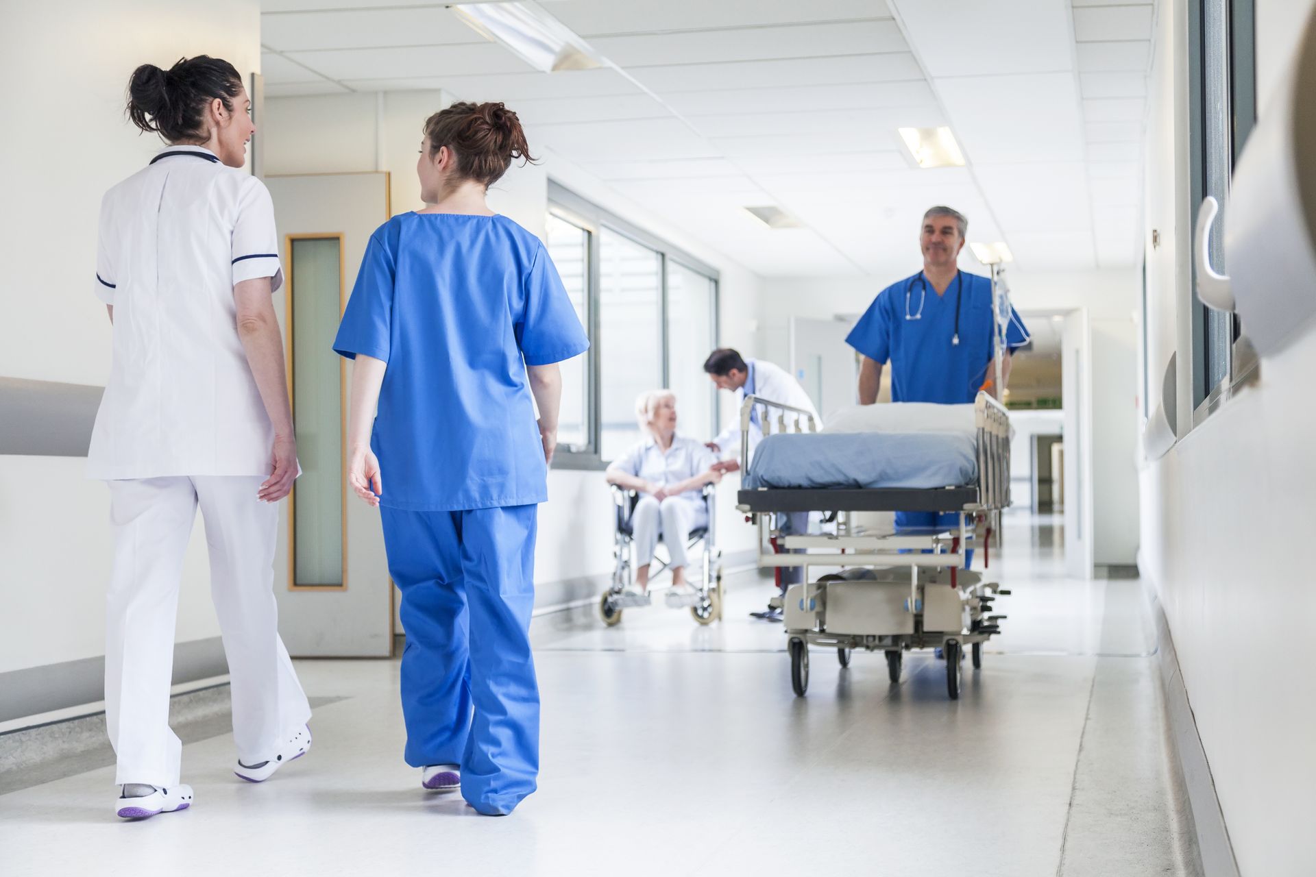 Hospital hallway with nurses, a doctor, and a patient in a wheelchair. A man pushes a gurney.